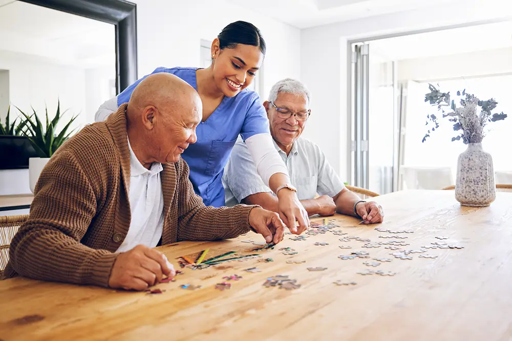 A caregiver at Sholom Senior Living helps two elderly men assemble a jigsaw puzzle at a wooden table in a bright, modern room filled with plants and a vase of flowers. A caregiver at Sholom Senior Living helps two elderly men assemble a jigsaw puzzle at a wooden table in a bright, modern room filled with plants and a vase of flowers.