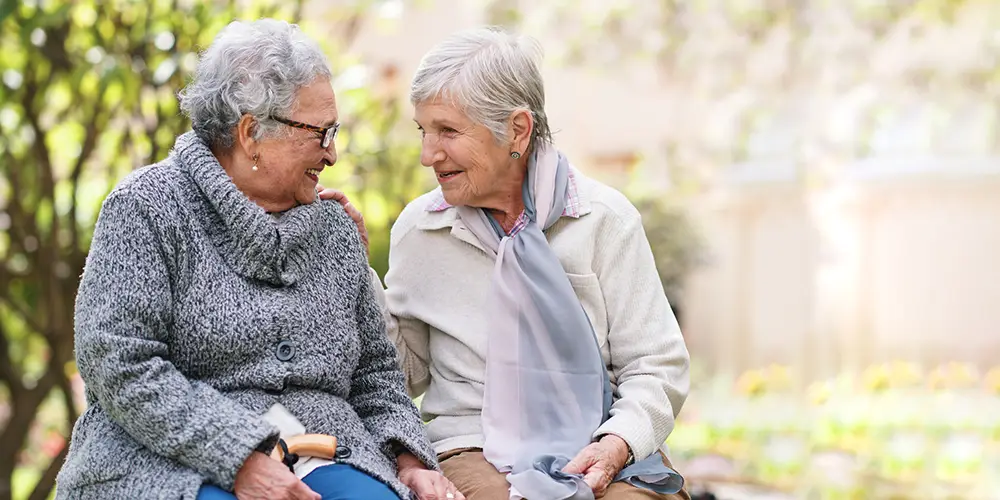 Two elderly women sit on a bench outdoors at Sholom Senior Living, facing each other and smiling, with greenery and flowers in the background. Two elderly women sit on a bench outdoors at Sholom Senior Living, facing each other and smiling, with greenery and flowers in the background.
