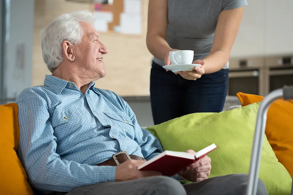 An older man sits on a couch at Sholom Senior Living, holding an open book while a person hands him a cup of coffee or tea. An older man sits on a couch at Sholom Senior Living, holding an open book while a person hands him a cup of coffee or tea.