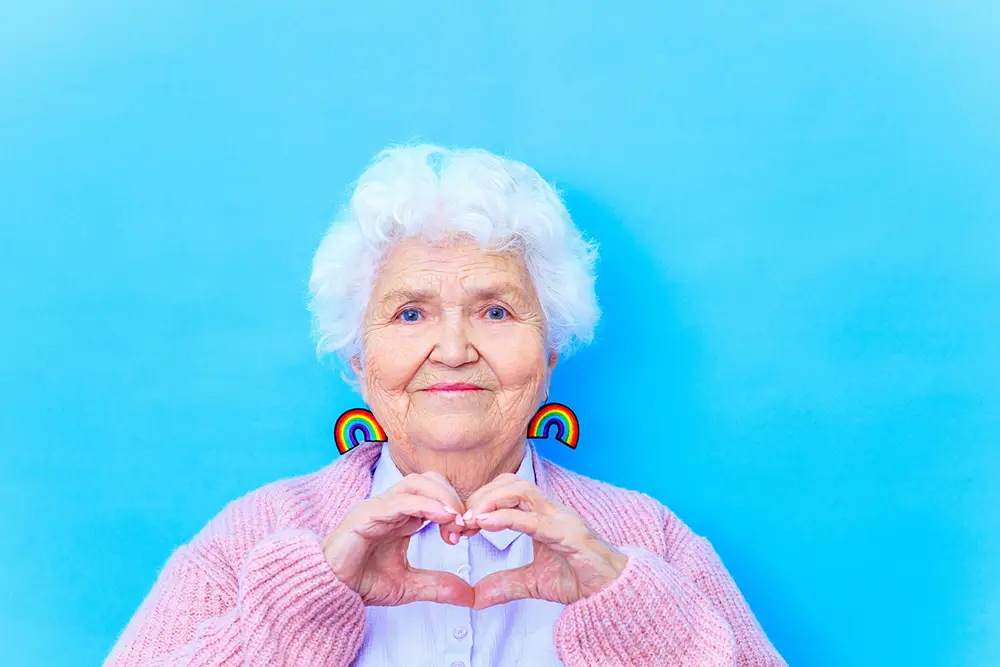 Elderly woman with white hair, wearing rainbow earrings and a pink cardigan, forms a heart shape with her hands against a bright blue background, embodying the warmth and community spirit of Sholom Senior Living. Elderly woman with white hair, wearing rainbow earrings and a pink cardigan, forms a heart shape with her hands against a bright blue background, embodying the warmth and community spirit of Sholom Senior Living.