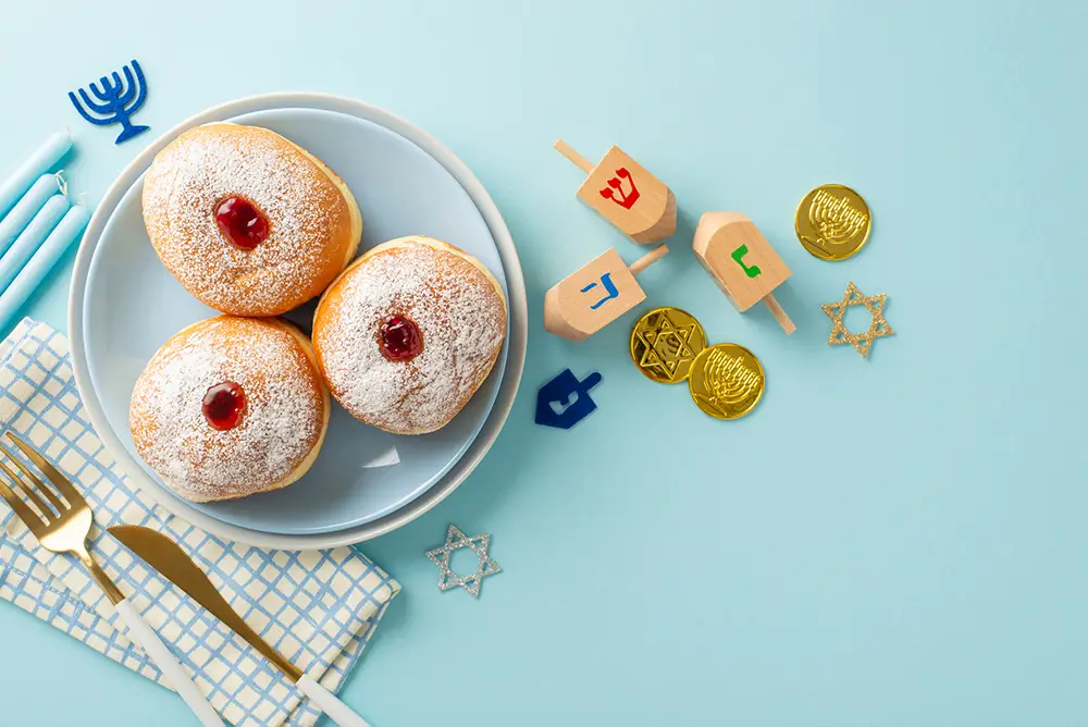 Three jelly-filled donuts on a plate with a napkin and cutlery, surrounded by Hanukkah candles, dreidels, chocolate coins, and Star of David decorations on a blue background at Sholom Senior Living. Three jelly-filled donuts on a plate with a napkin and cutlery, surrounded by Hanukkah candles, dreidels, chocolate coins, and Star of David decorations on a blue background at Sholom Senior Living.