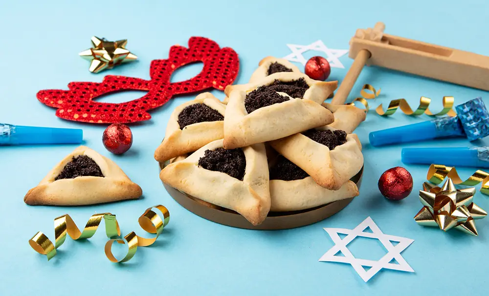 A plate of hamantaschen cookies surrounded by Purim decorations, including a mask, noisemaker, party blowers, star cutouts, and ribbons on a blue background at Sholom Senior Living. A plate of hamantaschen cookies surrounded by Purim decorations, including a mask, noisemaker, party blowers, star cutouts, and ribbons on a blue background at Sholom Senior Living.