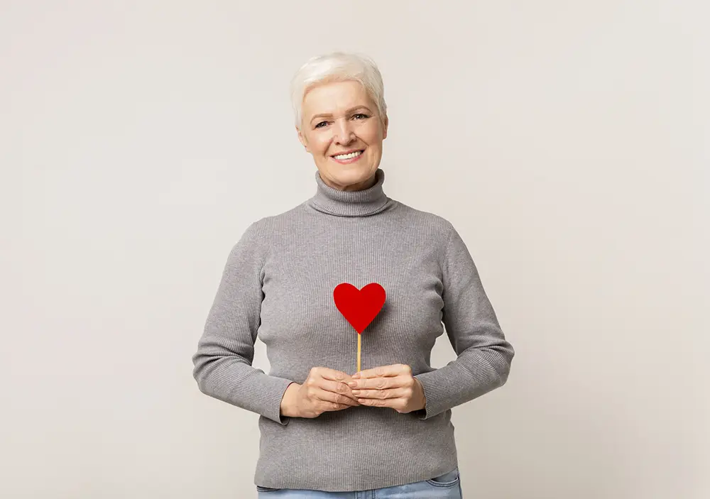 Older woman with short white hair, wearing a gray turtleneck sweater, smiles while holding a red glittery heart on a stick against a plain light background—capturing the warmth of Sholom Senior Living. Older woman with short white hair, wearing a gray turtleneck sweater, smiles while holding a red glittery heart on a stick against a plain light background—capturing the warmth of Sholom Senior Living.