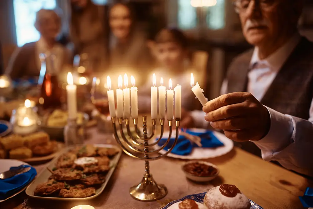 A person lights candles on a menorah at a Sholom Senior Living dinner table set with food, while others sit in the background. A person lights candles on a menorah at a Sholom Senior Living dinner table set with food, while others sit in the background.