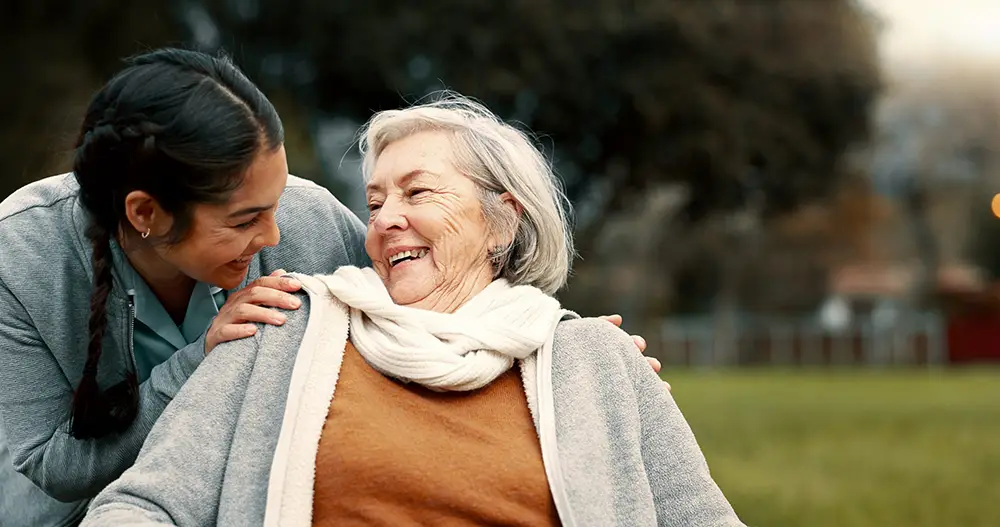A younger woman smiles and puts her hand on the shoulder of an older woman in a wheelchair. Both are outdoors on a grassy area, enjoying each other’s company at Sholom Senior Living. A younger woman smiles and puts her hand on the shoulder of an older woman in a wheelchair. Both are outdoors on a grassy area, enjoying each other’s company at Sholom Senior Living.