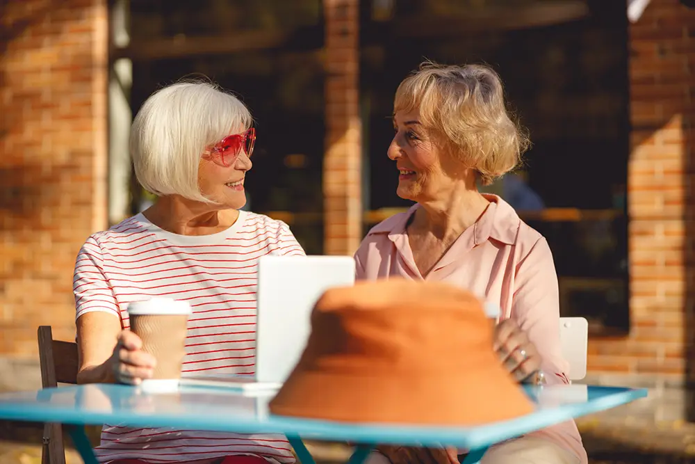 Two older women sit at an outdoor table at Sholom Senior Living, chatting and smiling over coffee and a tablet. A brown hat rests on the table, with a brick building in the background. Two older women sit at an outdoor table at Sholom Senior Living, chatting and smiling over coffee and a tablet. A brown hat rests on the table, with a brick building in the background.