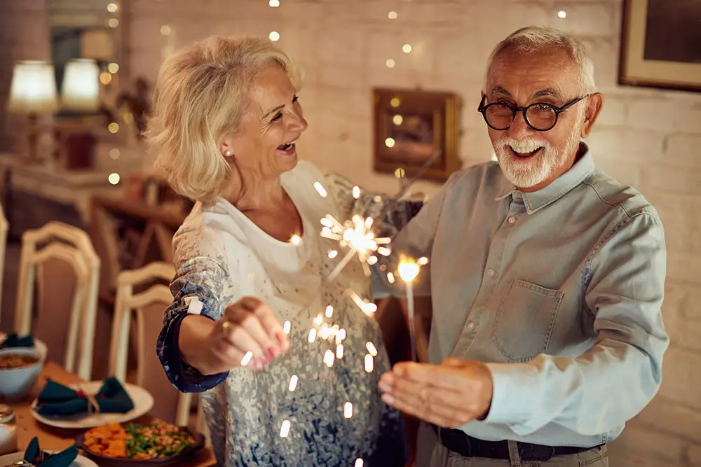 Older couple indoors holding sparklers and smiling, surrounded by a festive setting with a dining table in the background at Sholom Senior Living. Older couple indoors holding sparklers and smiling, surrounded by a festive setting with a dining table in the background at Sholom Senior Living.