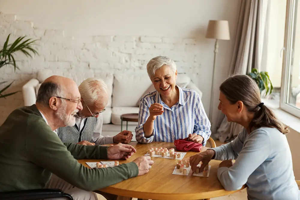 Four older adults sit around a table at Sholom Senior Living, playing a board game while smiling and laughing in a well-lit living room. Four older adults sit around a table at Sholom Senior Living, playing a board game while smiling and laughing in a well-lit living room.