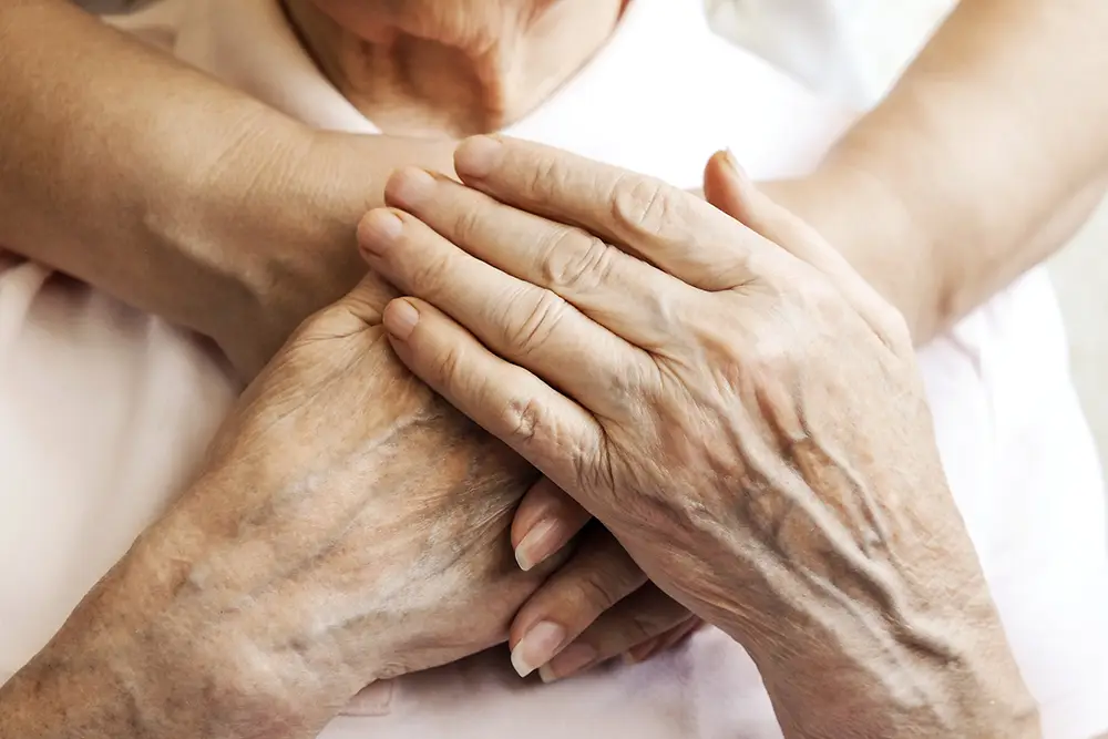 Close-up of an elderly person’s hands resting on their chest, with another person’s arms gently embracing them from behind at Sholom Senior Living. Close-up of an elderly person’s hands resting on their chest, with another person's arms gently embracing them from behind at Sholom Senior Living.