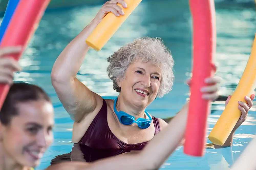 An older woman with short gray hair in a swimming pool at Sholom Senior Living, holding a yellow pool noodle and goggles around her neck, participates in a group aquatic exercise. An older woman with short gray hair in a swimming pool at Sholom Senior Living, holding a yellow pool noodle and goggles around her neck, participates in a group aquatic exercise.