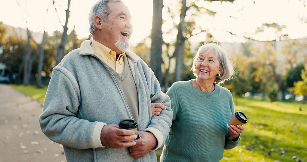 An older man and woman walk arm in arm through a park, smiling and holding coffee cups on a sunny day, enjoying the community spirit of Sholom Senior Living. An older man and woman walk arm in arm through a park, smiling and holding coffee cups on a sunny day, enjoying the community spirit of Sholom Senior Living.