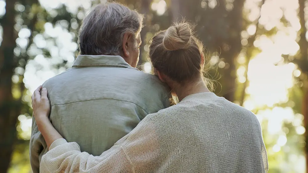 Two people are seen from behind; one has an arm around the other, enjoying the outdoors at Sholom Senior Living with sunlight and trees in the background. Two people are seen from behind; one has an arm around the other, enjoying the outdoors at Sholom Senior Living with sunlight and trees in the background.