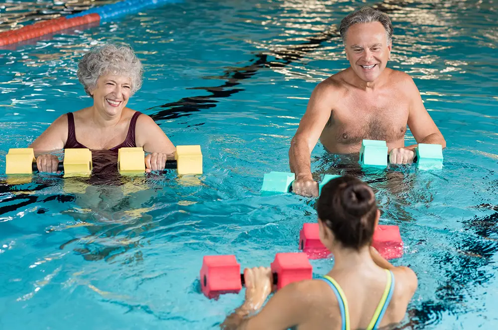 Two older adults at Sholom Senior Living take part in a water exercise class, using foam dumbbells and following the guidance of an instructor in a swimming pool. Two older adults at Sholom Senior Living take part in a water exercise class, using foam dumbbells and following the guidance of an instructor in a swimming pool.