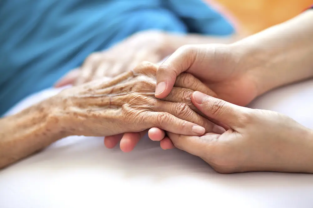 A younger person gently holds the hand of an elderly person lying in bed at Sholom Senior Living, showing care and support. A younger person gently holds the hand of an elderly person lying in bed at Sholom Senior Living, showing care and support.
