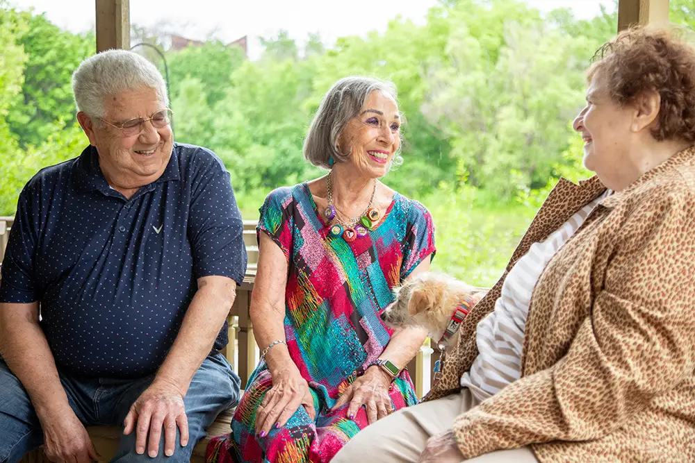 Three older adults sit together on a porch at Sholom Senior Living, smiling and talking; a small dog rests on the lap of the woman in the colorful dress. Greenery is visible in the background.