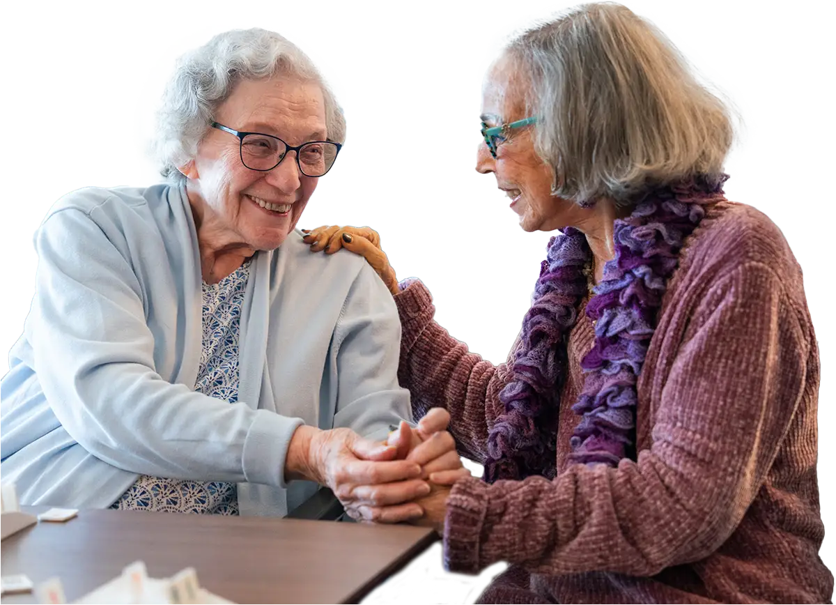 Two elderly women sit at a table at Sholom Senior Living, smiling and holding hands. One wears a blue sweater, the other a purple scarf, as they engage in a friendly conversation. Two elderly women sit at a table at Sholom Senior Living, smiling and holding hands. One wears a blue sweater, the other a purple scarf, as they engage in a friendly conversation.